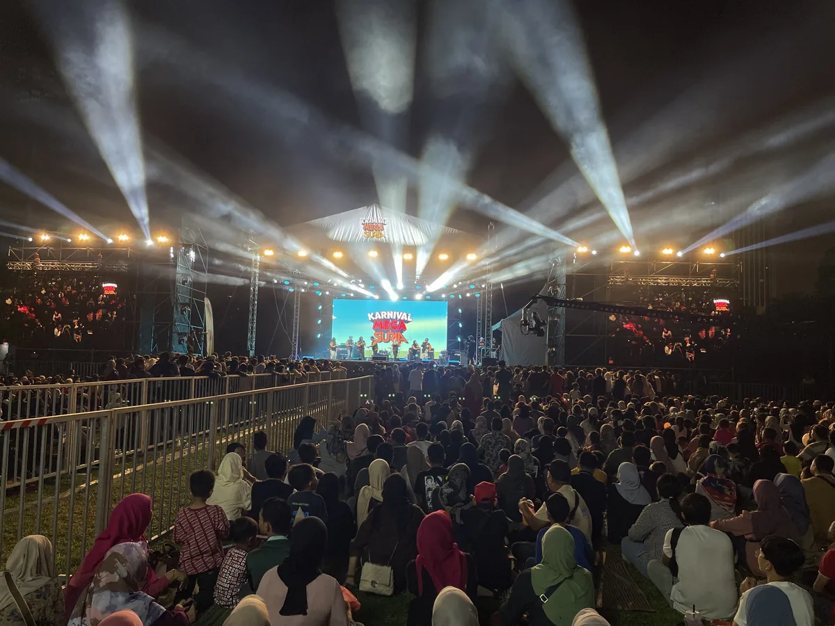 Night crowd facing a lit stage with white beams and a central LED screen